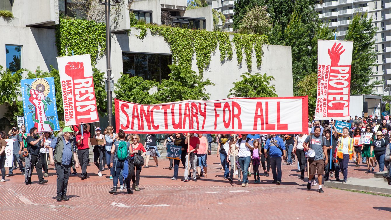 protestors march with a white banner with red text reading "sanctuary for all"