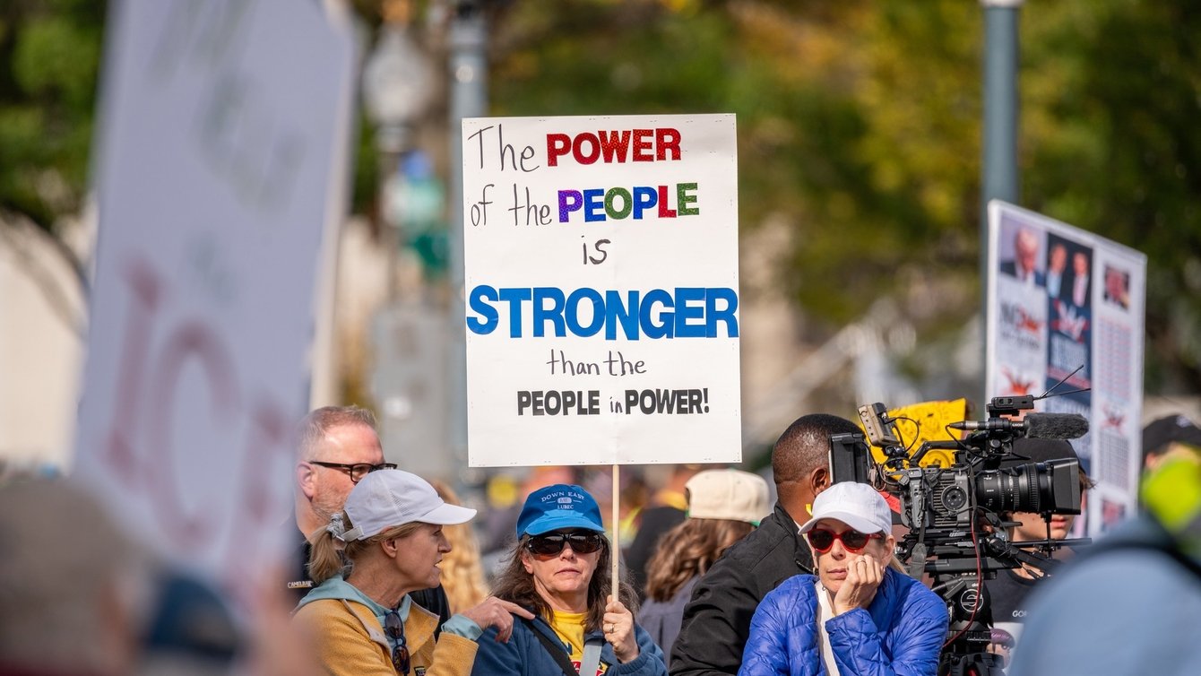 a group of people behind a barrier. The person in the center holds a sign that reads "the power of the people is stronger than the people in power."