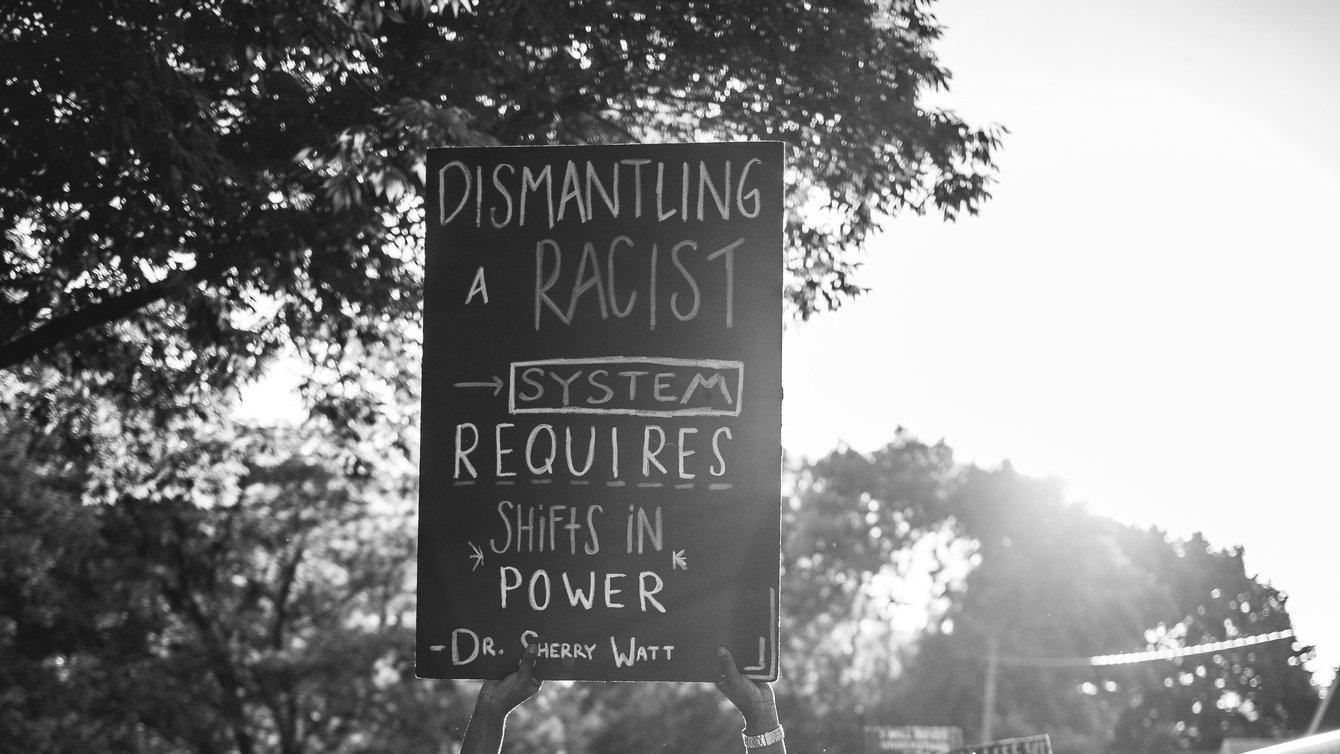 a person holding a sign that reads "dismantling a racist system requires shifts in power - Dr. Sherry Watt"