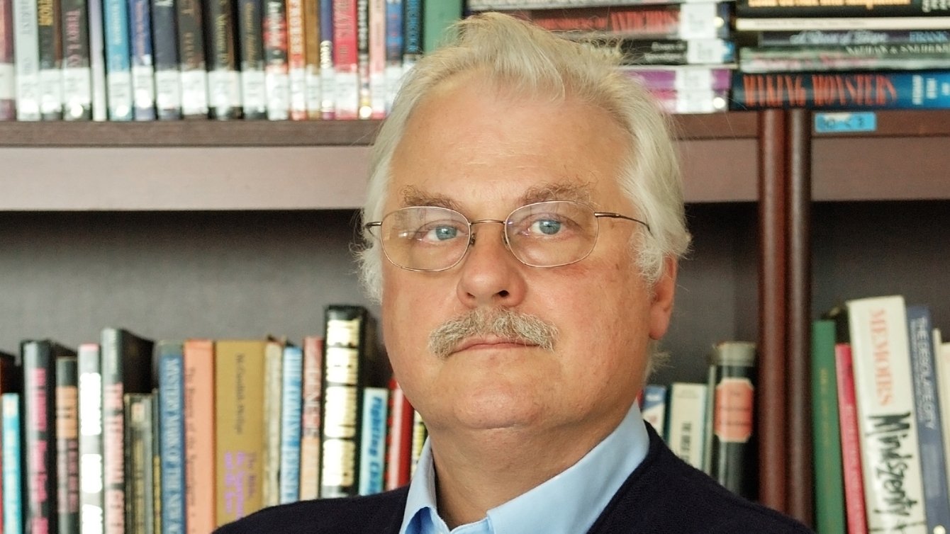 a man with white hair, glasses, and a moustache stands in front of a book case