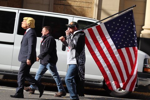 Three men walking; one wears a Trump mask and one carries an American flag