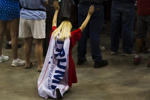 A white woman with blond hair, kneeling with her arms in the air, a Trump flag tied around her neck and hanging down her back