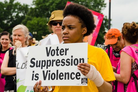 A Black person in a yellow T-shirt holding a sign that says "voter suppression is violence"