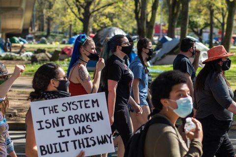 Protesters wearing masks. One protester holding a sign that says "The system isn't broken it was built this way."