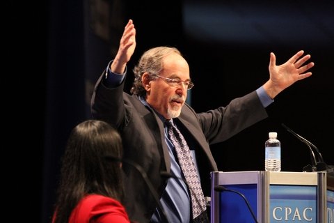 David Horowitz, a white man with his hands raised in the air, in front of a mic stand.