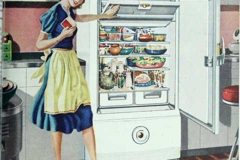 A white woman in a blue dress and a yellow apron, standing in front of an open fridge full of food.