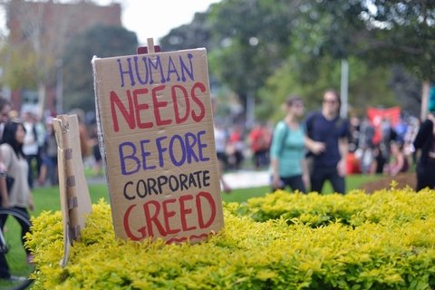 A cardboard sign that says Human Needs Before Corporate Greed