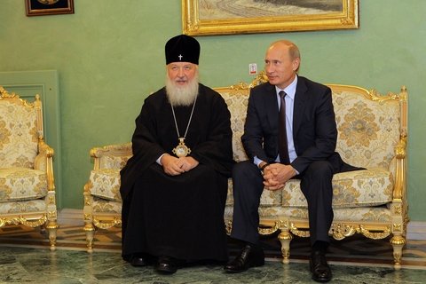 A man in a black robe and a black hate with a cross on it sitting next to Vladimir Putin on a golden couch.