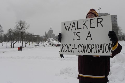 A man in winter clothes holding a board that says "walker is a koch-conspirator"