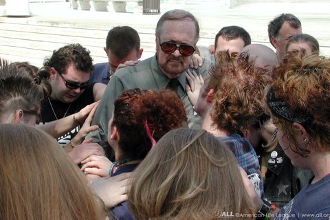 A white man in the center of a circle, with other people bowing their heads and placing a hand on his shoulder and arms.
