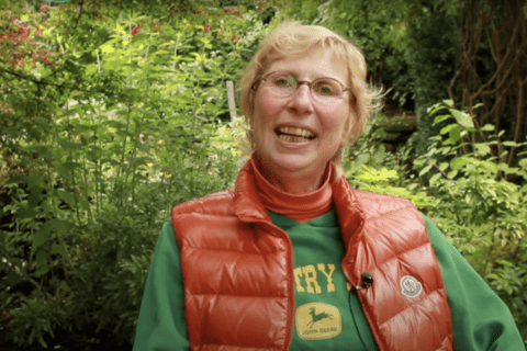 A White woman with short blond hair wearing a green t-shirt and orange sleeveless jacket sitting in front of some bushes.