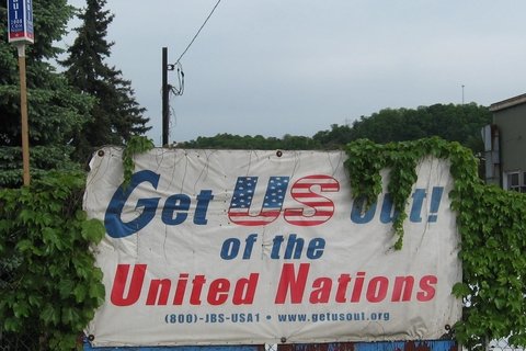A sign covered by plants. The sign reads "Get US out! of the United Nations (888) JBS USA1"