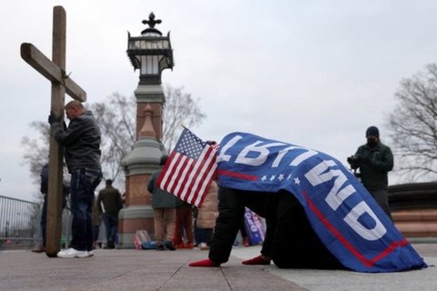 A man wearing a Trump flag bowing in front of a cross.
