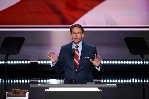A white man in the suit standing on a podium talking into a mic