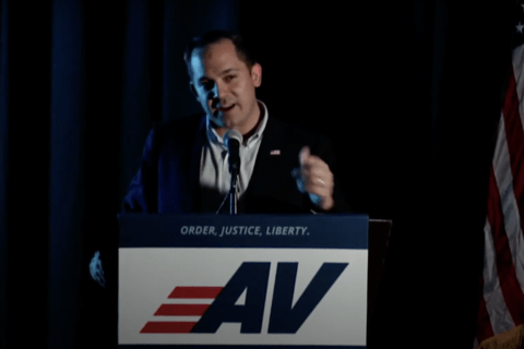 A man in blue suit standing at a podium with the letters AV on it speaking into a mic with two American flags behind him.