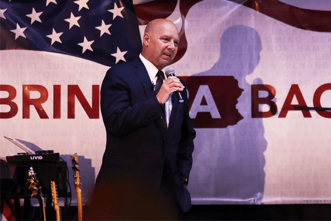 A bald White man on a stage, speaking into a mic