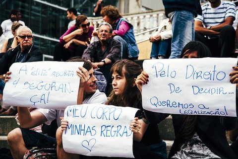People sitting on stairs. Three people hold signs in Portugese, written with blue ink on white paper.