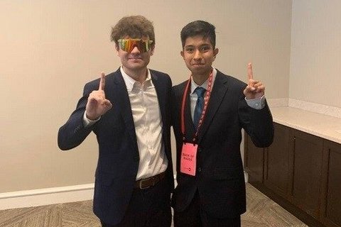 Two men in suits without ties holding up one finger.