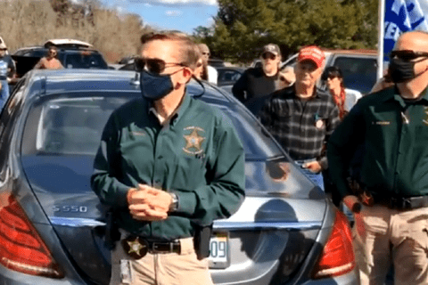A white man in a mask holding his writs, wearing sunglasses and a greenshirt with the homeland security logo on it.