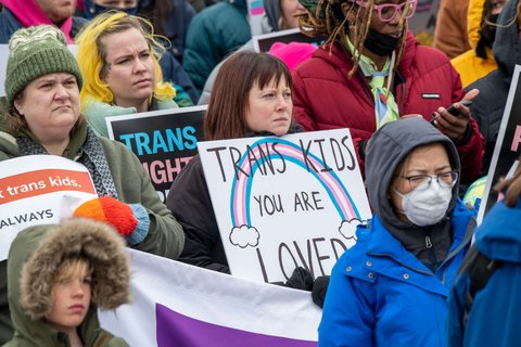 People in jackets and hats, holding signs supporting trans rights.