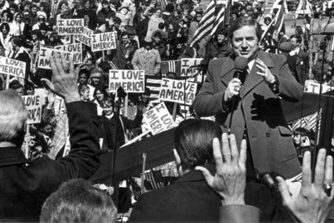 Jerry Falwell, a white man wearing a suit coat, speaks in front of a crowd holding signs reading "I love America."