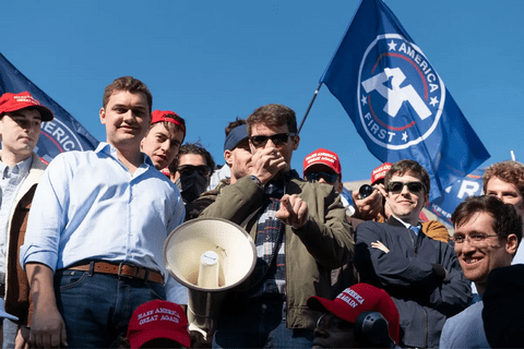 Nick Fuentes and other young men wearing sun glasses and hats carrying an America First flag.