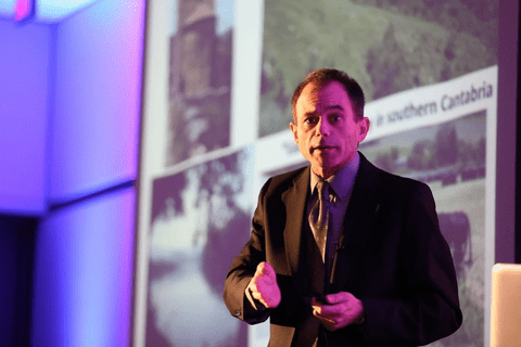 A man in a brown suit looking into the camera in the middle of a speech.