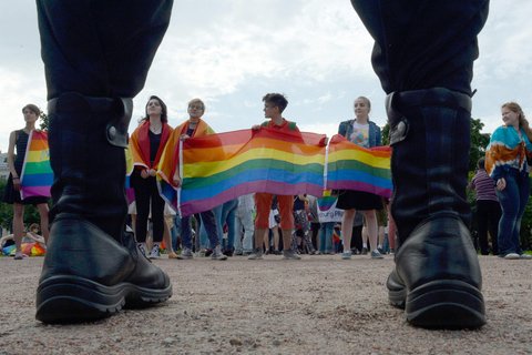 Shoes standing opposite a group of people holding a rainbow flag.