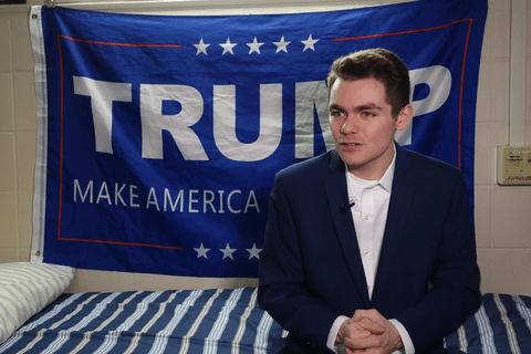 Nick Fuentes, a White man with brown hair sitting in front of Trump flag.
