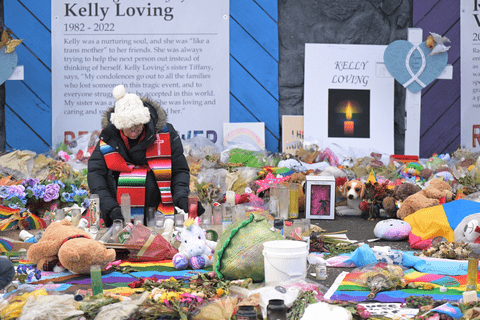a person in a hat and scarf kneeling at the memorial outside Club Q