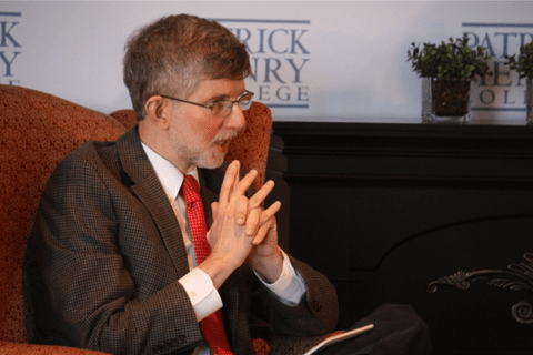A white man wearing a brown suit sitting in an orange arm chair.