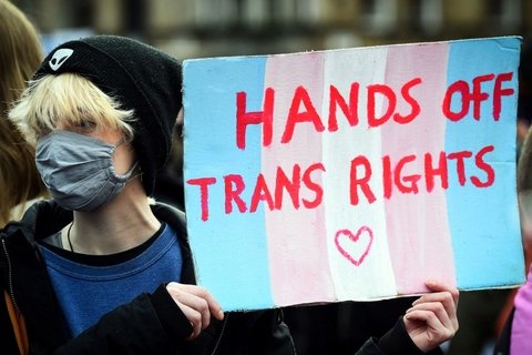 A person with blonde hair wearing a black winter hate holding a sign that says "Hands Off Trans Rights"