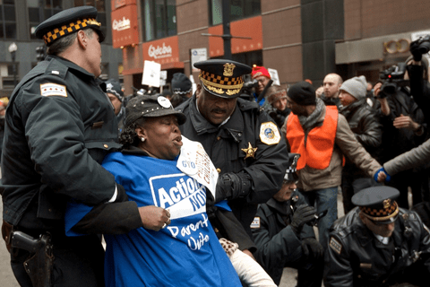 a black woman in a blue t-shirt and black hat being dragged away by policemen.