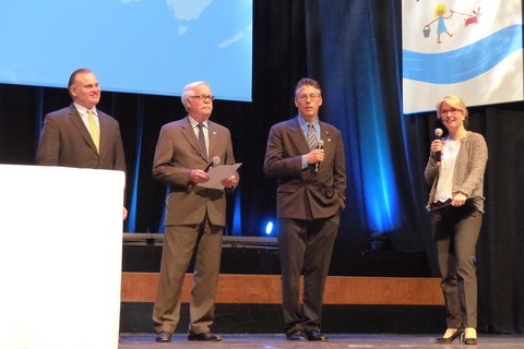 Three men in suits and a woman holding a mic.