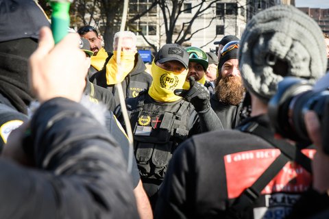 A Proud Boy with a neck warmer covering his mouth points mid-speech at the camera