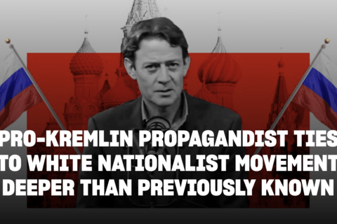 A man in a suit standing in front of the Kremlin with Russian flags on either side.