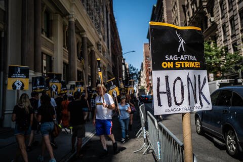 Protesters in the background. The Foreground has a sign that says "SAF-Aftra On Strike! Honk"