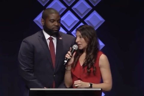 A Black man in a suit and a White woman in a red dress speaking at a podium