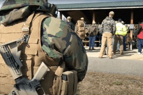 A man in camouflage in the foreground with a gun on his shoulder.