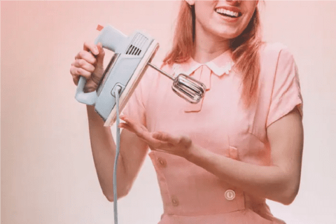 a white woman in a pink dress holding an electric mixer.