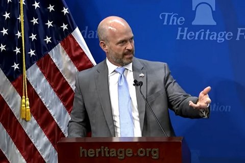 A bald man in a grey suit speaking into a mic