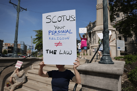 a protestor holding a sign that says "SCOTUS: Your Personal Religion does not equal the law"