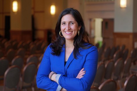 Smiling woman with long brown hair, gold hoop earrings, and a blue blazer, arms crossed
