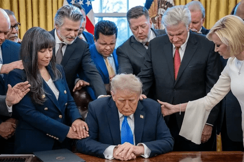 President Trump surrounded by people, bowing their heads and placing their hands on him.