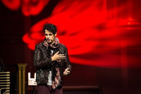 A man in a black jacket speaks in front of a red blurred background 