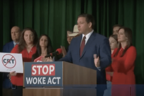 a man in a blue suit standing behind a podium with a sign that says "stop woke act"