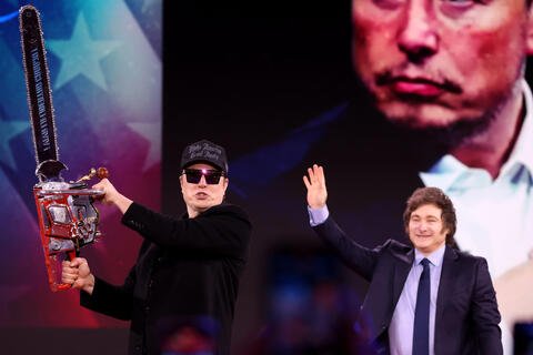 Two men standing in front of projector image. One man is waving at the crowd, the other man is holding a chainsaw