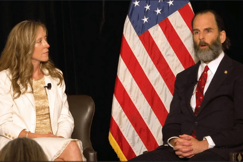two people sitting on a stage with an American flag behind them.