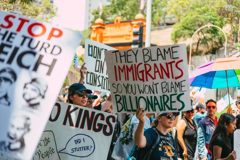 a person holding a sign that says "they blame the immigrants so you won't blame billionaires"