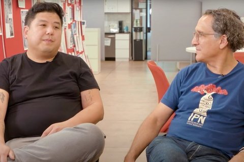 Two men sitting in chairs, facing the camera but looking at each other.